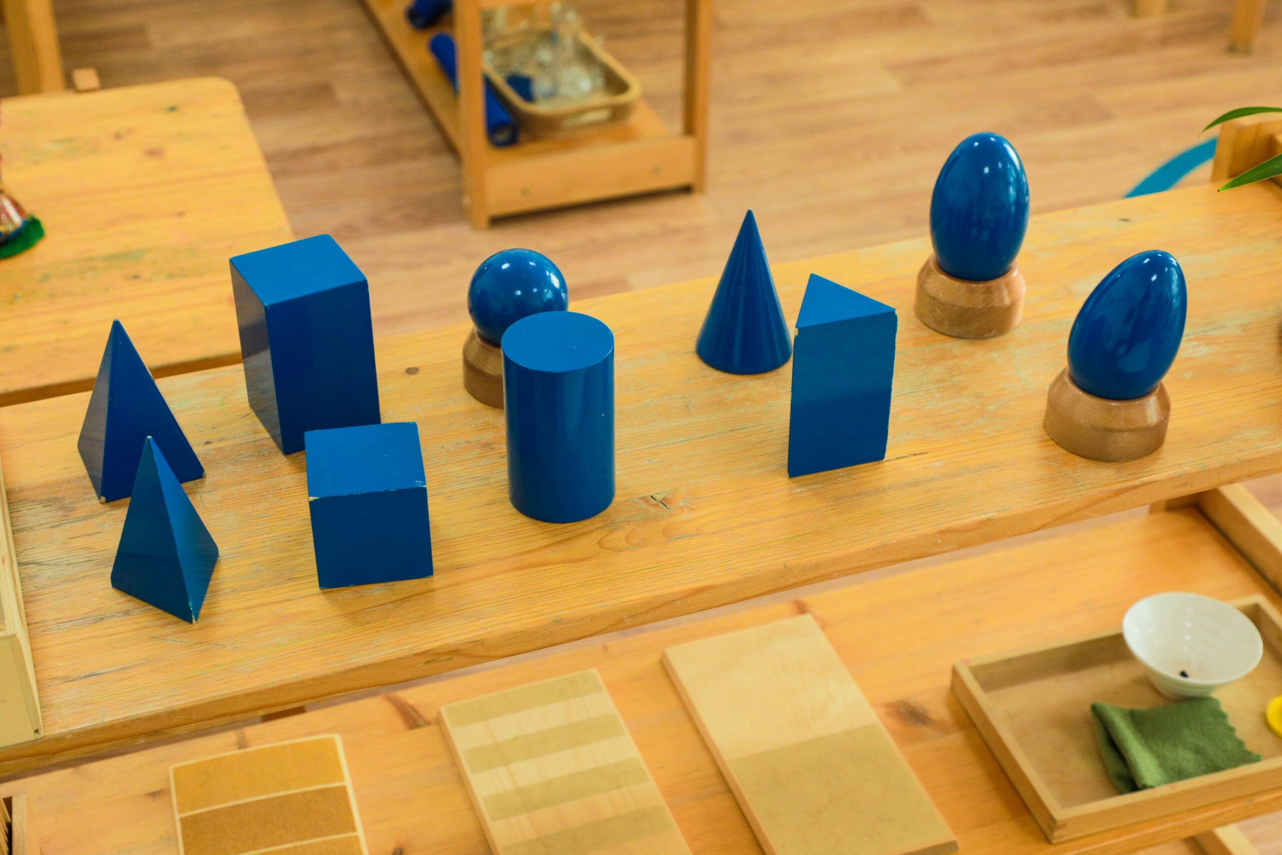 Sólidos geométricos azuis sobre mesa infantil: esfera, cilindro, cone e prismas.